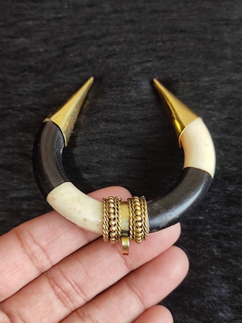 May include: A handcrafted, U-shaped pendant necklace. The pendant features a curved design with black and cream sections, accented with gold-colored metal tips and a central decorative element. The necklace is designed to be worn as a statement piece.