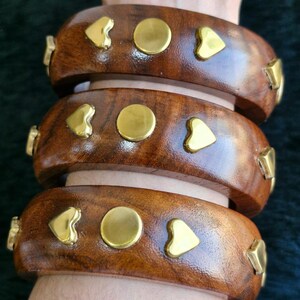 May include: Three brown wooden bangles with gold-colored heart, circle, and triangle accents. The bangles are stacked on top of each other, showcasing the decorative details and the rich wood grain. The jewelry is designed to be worn on the wrist.