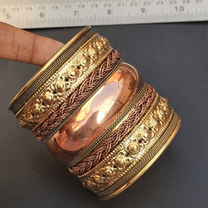 Op de afbeelding: Een gouden en koperen armband met ingewikkelde geweven en gegraveerde details. De armband is gemaakt van meerdere lagen metaal, waardoor een textuur en gelaagdheid ontstaat.