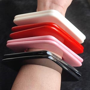 May include: A stack of square-shaped bangles in white, red, pink, and black. The bangles are stacked on a wrist, showcasing their glossy finish and vibrant colors. The bracelets have a smooth, curved design.