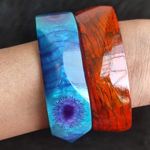 May include: Two rectangular bangles. One is a vibrant blue with a central purple design, the other is a rich red-orange with dark wood grain patterns. The bangles are worn on a wrist, showcasing their colors and textures.