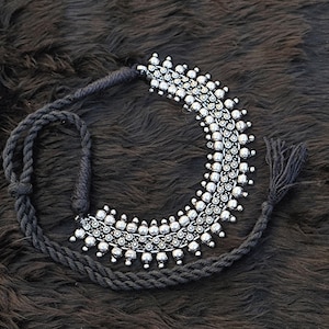 Op de afbeelding: Een zilveren chokerketting met een kralenontwerp en een zwarte koordsluiting. De ketting heeft aan één uiteinde een kwast.