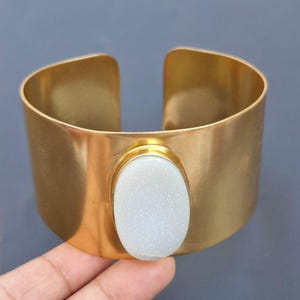 May include: A gold cuff bracelet with a white oval-shaped gemstone set in the centre.