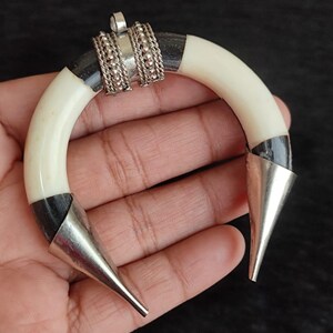 May include: A handcrafted pendant with a curved, ivory-colored body, accented with black sections and silver metal cone-shaped tips. The center features ornate silver detailing. This unique piece is likely a statement jewelry item.