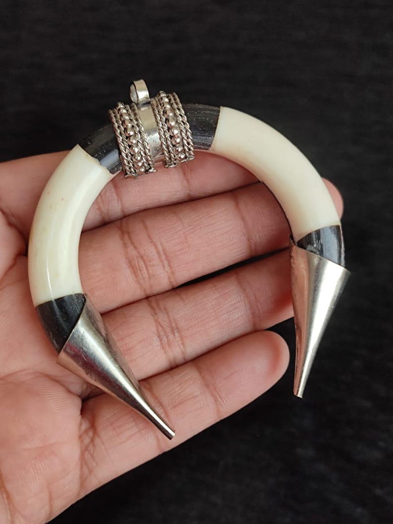 May include: A handcrafted pendant with a curved, ivory-colored body, accented with black sections and silver metal cone-shaped tips. The center features ornate silver detailing. This unique piece is likely a statement jewelry item.
