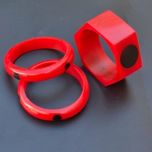 May include: Three red plastic bangles with black polka dots. The bangles are stacked on top of each other. The bangle on the bottom is a wider, rectangular shape.