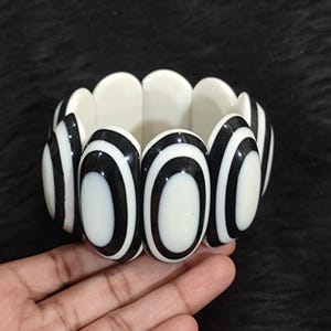 May include: A stretch bracelet composed of repeating oval-shaped links. Each link features a high-contrast design with a solid white center surrounded by a thick black border. The bracelet has a flexible construction that allows it to expand.