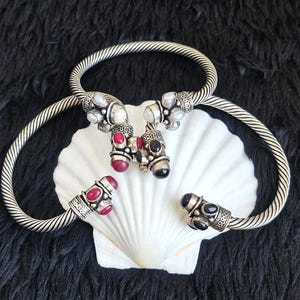 Puede incluir: Dos brazaletes abiertos de estilo cable plateado. Los extremos están decorados con pequeñas piedras preciosas redondas y pulidas en tonos rojo, negro y blanco, dispuestas en grupos sobre los engastes de metal.