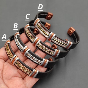 May include: Four leather bracelets with copper and silver accents. The bracelets are stacked on top of each other, with the top bracelet being the most visible. The bracelets are all black leather with a braided design. The copper and silver accents are in the form of small beads and strips.