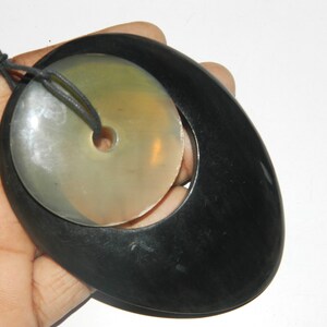 May include: A black oval pendant with a round, white, and green stone inside. The pendant has a hole in the center of the stone and is attached to a black cord.