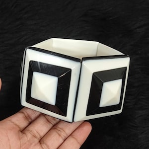 Puede incluir: Una pulsera rígida ancha con un diseño geométrico de segmentos alternos en forma de pirámide cuadrada de color blanco y negro. La pulsera tiene una estructura gruesa y rígida con un acabado liso y pulido, mostrando un patrón de alto contraste.