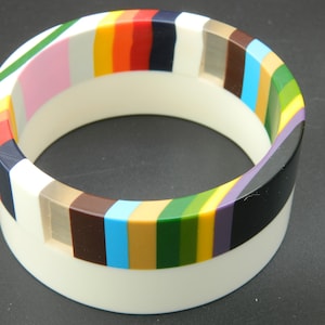 Op de afbeelding: Een witte bangle-armband met een kleurrijk gestreept ontwerp. De strepen zijn in verschillende kleuren, waaronder roze, rood, oranje, geel, groen, blauw, paars en zwart.