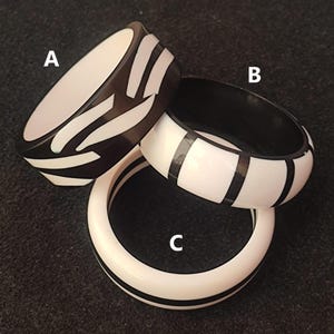 May include: Three black and white bangles. Bangle A is black with white geometric shapes. Bangle B is white with black stripes. Bangle C is white with black stripes. The bangles are made of a glossy material.