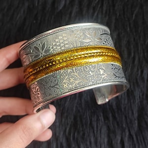 May include: A wide cuff bracelet featuring a silver-toned metal base with intricate floral and vine engravings. The centre of the cuff is accented by two horizontal, textured gold-toned bands that run parallel across the middle of the piece.