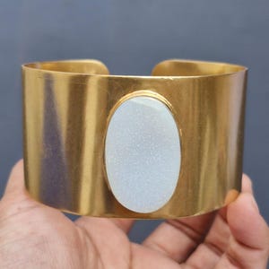 May include: A gold metal cuff bracelet with a white oval-shaped stone set in the centre.