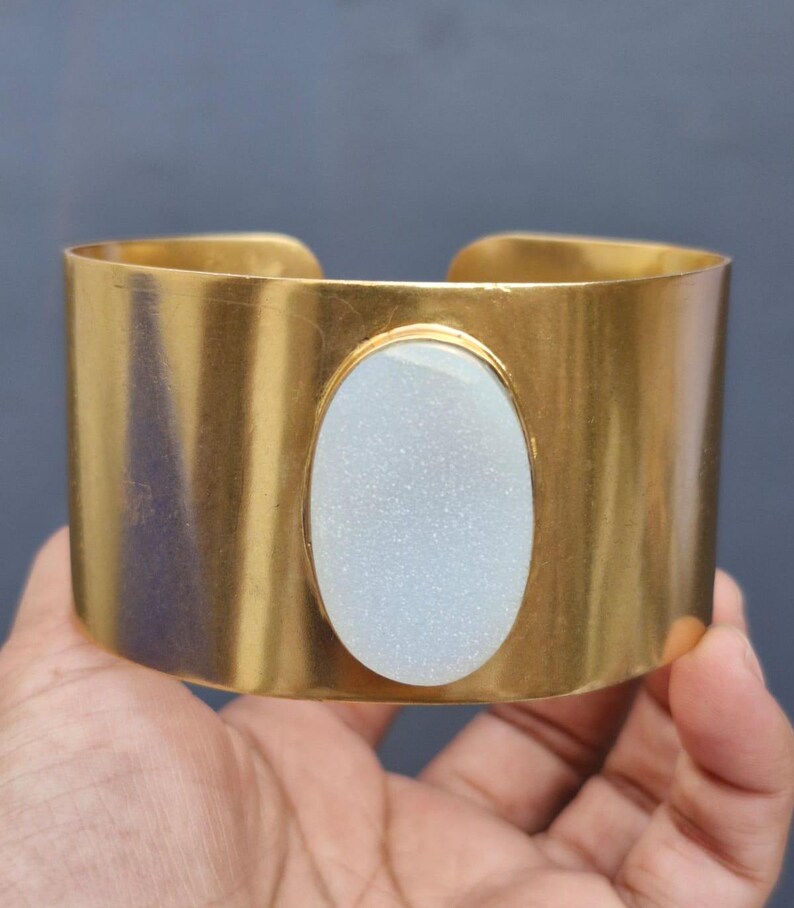 May include: A gold metal cuff bracelet with a white oval-shaped stone set in the centre.