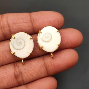 Könnte beinhalten: Zwei weiße Muschelkabochen mit goldfarbenen Zargen und Sprungringen. Die Muscheln haben ein Spiralmuster.