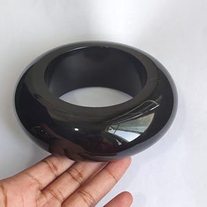 May include: A thick, solid black bangle bracelet with a smooth, high-gloss finish. The circular accessory features a wide, rounded profile and a reflective surface that catches light, creating a polished appearance throughout the entire piece.