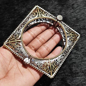 May include: A silver and gold square bangle bracelet with intricate filigree designs. The bracelet has a hinged clasp and is decorated with small beads around the edge.