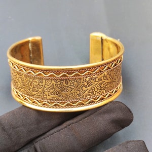 Könnte beinhalten: Ein goldfarbenes Metall-Armband mit einem komplizierten Blumenmuster. Das Armband hat eine strukturierte Oberfläche und eine leicht gebogene Form.
