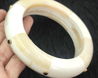 May include: A cream-colored bangle bracelet with a segmented design. The bracelet features alternating sections of white and light beige. Small gold-colored accents with black stones are evenly spaced around the bracelet.