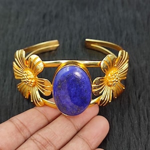 May include: Gold-coloured cuff bracelet with a large, oval, deep blue gemstone. The bracelet has floral accents on either side of the gemstone. The cuff is open at the back. The bracelet is displayed against a dark background.