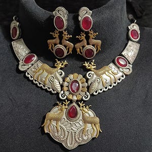 Könnte beinhalten: Ein silberfarbenes Halsketten- und Ohrring-Set mit roten Edelsteinakzenten. Die Halskette hat einen zentralen Anhänger mit Hirschfiguren und ein passendes Paar Ohrringe. Der Schmuck hat goldfarbene Hirschfiguren und dekorative Silberarbeiten.