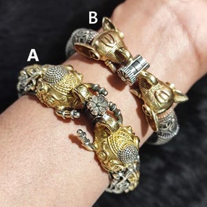 Puede incluir: Dos pulseras de plata y oro con diseños de cabeza de elefante. Las pulseras están en la muñeca de una persona. La pulsera de arriba está etiquetada con "B" y la pulsera de abajo está etiquetada con "A".