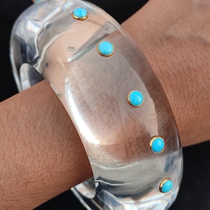 May include: A clear, wide bangle bracelet with turquoise-colored stones set in gold-colored bezels. The bracelet is worn on a wrist, showcasing its smooth, transparent design and the vibrant contrast of the blue stones against the gold and clear material.