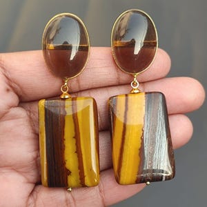 May include: A pair of gold tone earrings with a brown and yellow striped rectangular stone and a clear oval stone.