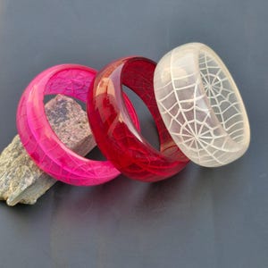 Può includere: Tre braccialetti colorati. Un braccialetto rosa acceso con una superficie strutturata, un braccialetto rosso intenso e un braccialetto bianco traslucido con un disegno a ragnatela. I braccialetti sono esposti su una superficie grigio scuro.