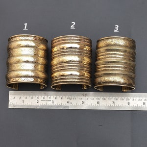 Peut inclure: Trois bracelets en métal doré avec des motifs gravés complexes. Les bracelets sont empilés sur une règle, avec les numéros 1, 2 et 3 étiquetés au-dessus de chaque bracelet.