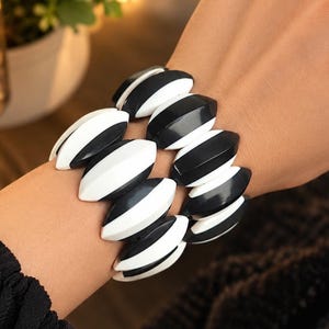 Puede incluir: Una pulsera elástica compuesta por múltiples cuentas lisas en forma de marquesa en colores alternos blanco y negro. Las cuentas están dispuestas en un patrón repetitivo, creando un diseño audaz y de alto contraste alrededor de la muñeca.