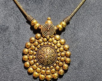 May include: A gold-colored necklace with a circular pendant. The pendant features a spiral design at the top and a detailed, textured pattern of small spheres and concentric circles. The necklace chain is a thin, gold-toned metal.