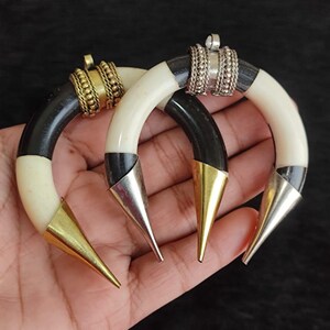 May include: Two horn-shaped earrings with a black and cream color scheme. The earrings have gold and silver metal accents, including cone-shaped tips and decorative rings. The earrings are displayed against a dark background.