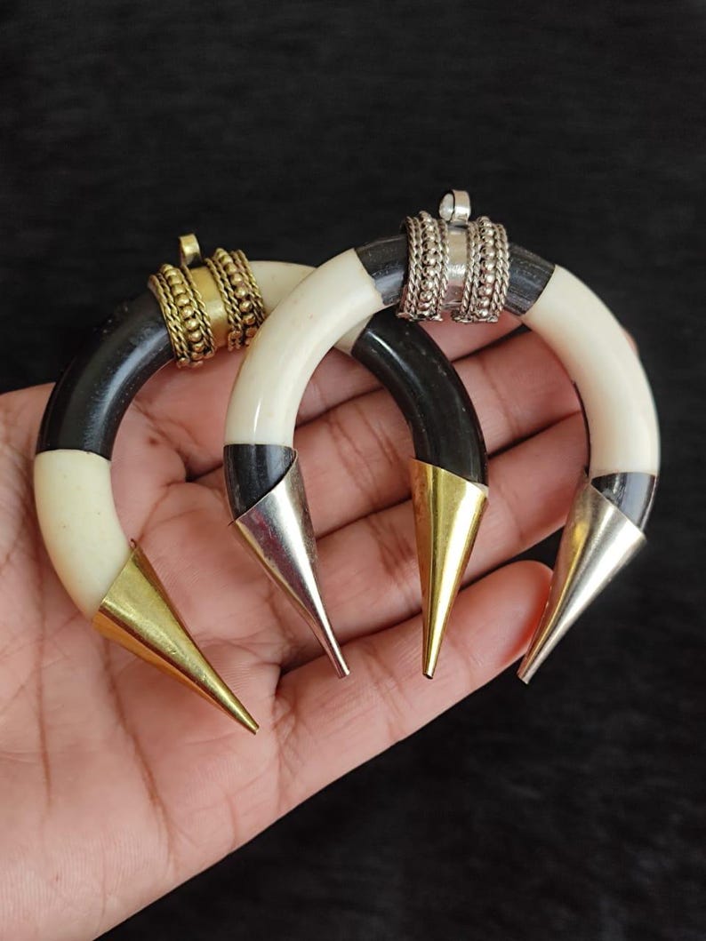 May include: Two horn-shaped earrings with a black and cream color scheme. The earrings have gold and silver metal accents, including cone-shaped tips and decorative rings. The earrings are displayed against a dark background.