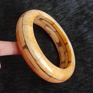May include: A cream-coloured bangle bracelet with a mosaic-like design. The bracelet is made of rectangular pieces in shades of cream and brown. The bracelet is a circular shape and appears to be made of a hard material.