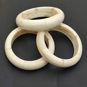 Op de afbeelding: Drie ivoren armbanden gestapeld op elkaar. De armbanden zijn gemaakt van een materiaal dat lijkt op bot of schelp en hebben een licht getextureerd oppervlak.