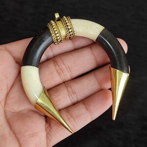 May include: A crescent-shaped pendant with a black, cream, and gold color scheme. The pendant features a curved design with black and cream segments, connected by gold-colored metal accents. The ends are capped with pointed gold metal cones.