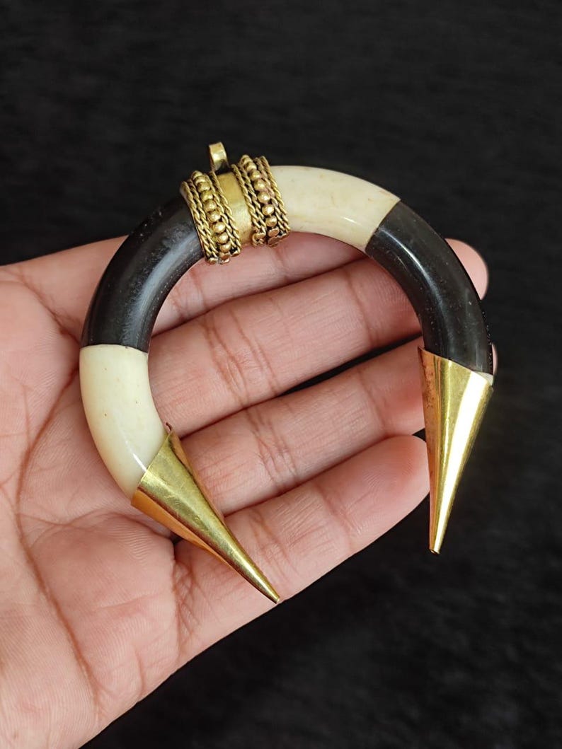 May include: A crescent-shaped pendant with a black, cream, and gold color scheme. The pendant features a curved design with black and cream segments, connected by gold-colored metal accents. The ends are capped with pointed gold metal cones.