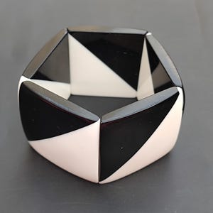 Peut inclure: Un bracelet géométrique noir et blanc en plastique. Le bracelet est composé de formes triangulaires disposées selon un motif répétitif.