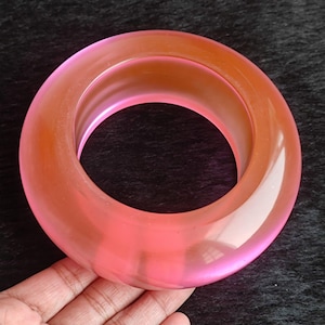 May include: A translucent, pink and orange bangle bracelet. The circular bracelet has a smooth, glossy finish. The colour transitions from a vibrant pink to a warm orange hue. The bracelet is shown against a dark background.