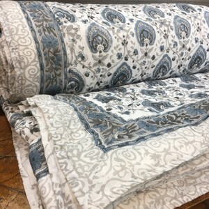 May include: A white cotton quilt with a blue and grey paisley print. The quilt is folded in half and the edges are visible.