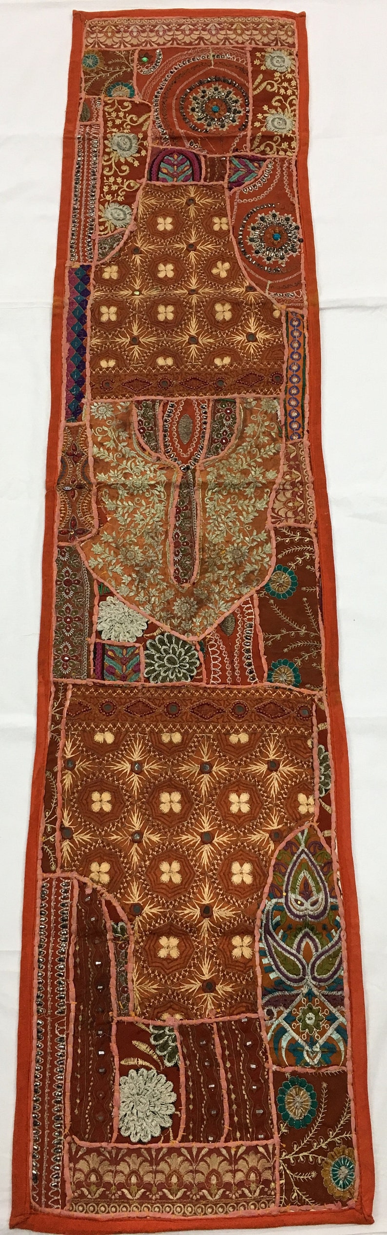 May include: A long, rectangular tapestry with a patchwork design. The tapestry is made of various fabrics in shades of brown, orange, pink, and green. It features intricate embroidery, sequins, and beads. The tapestry has a fringe border.