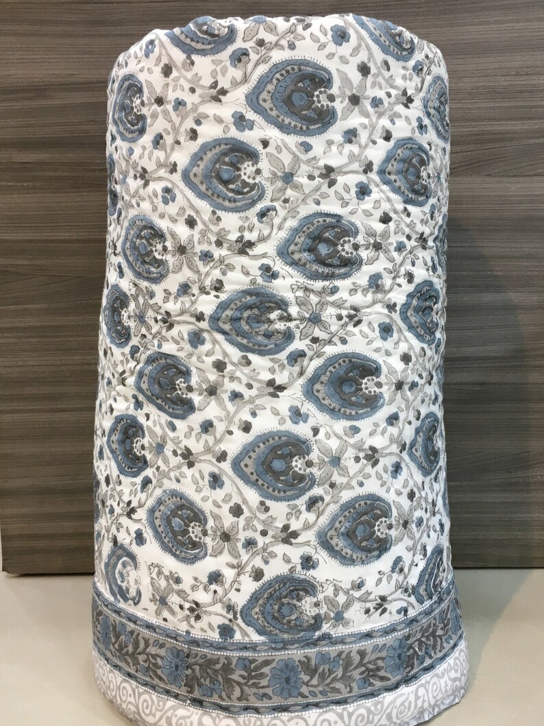 May include: A white cotton quilt with a blue and grey paisley pattern. The quilt is rolled up and has a border with a floral design.