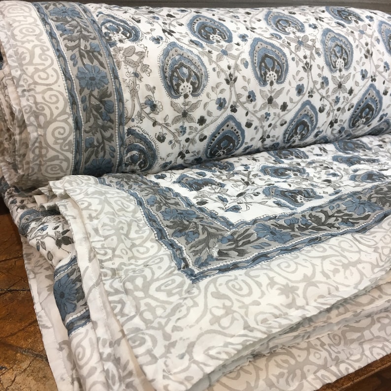 May include: A white cotton quilt with a blue and grey paisley print. The quilt is folded in half and the edges are visible.