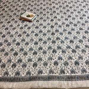 May include: A white cotton quilt with a blue and brown floral pattern. The quilt has a border with a repeating leaf design.