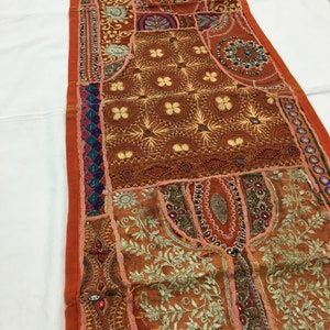 May include: A long, rectangular tapestry with a patchwork design. The tapestry is made of various fabrics in shades of orange, brown, and gold. It features intricate embroidery, sequins, and beads.