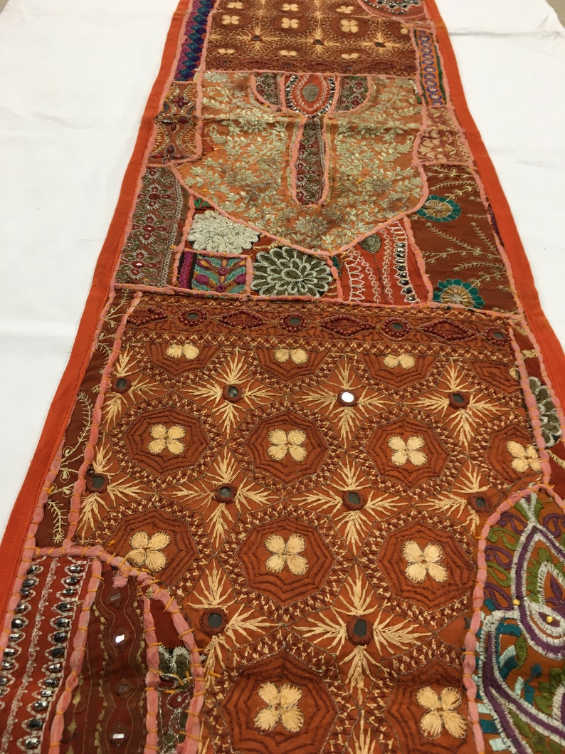 May include: A long, rectangular tapestry with a patchwork design. The tapestry is made of various fabrics in shades of brown, orange, pink, and green. It features intricate embroidery, sequins, and small mirrors.