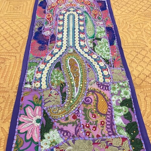 May include: A vibrant, rectangular textile with a patchwork design. The central motif features a paisley shape with intricate embroidery and beadwork. The color palette includes purple, pink, green, and gold, with a deep purple border.